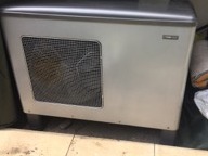 Biała jednostka centralna systemu wentylacji mechanicznej z odzyskiem ciepła, widoczna metalowa kratka wentylacyjna, szara obudowa, na podłodze widoczne fragmenty innych przedmiotów.