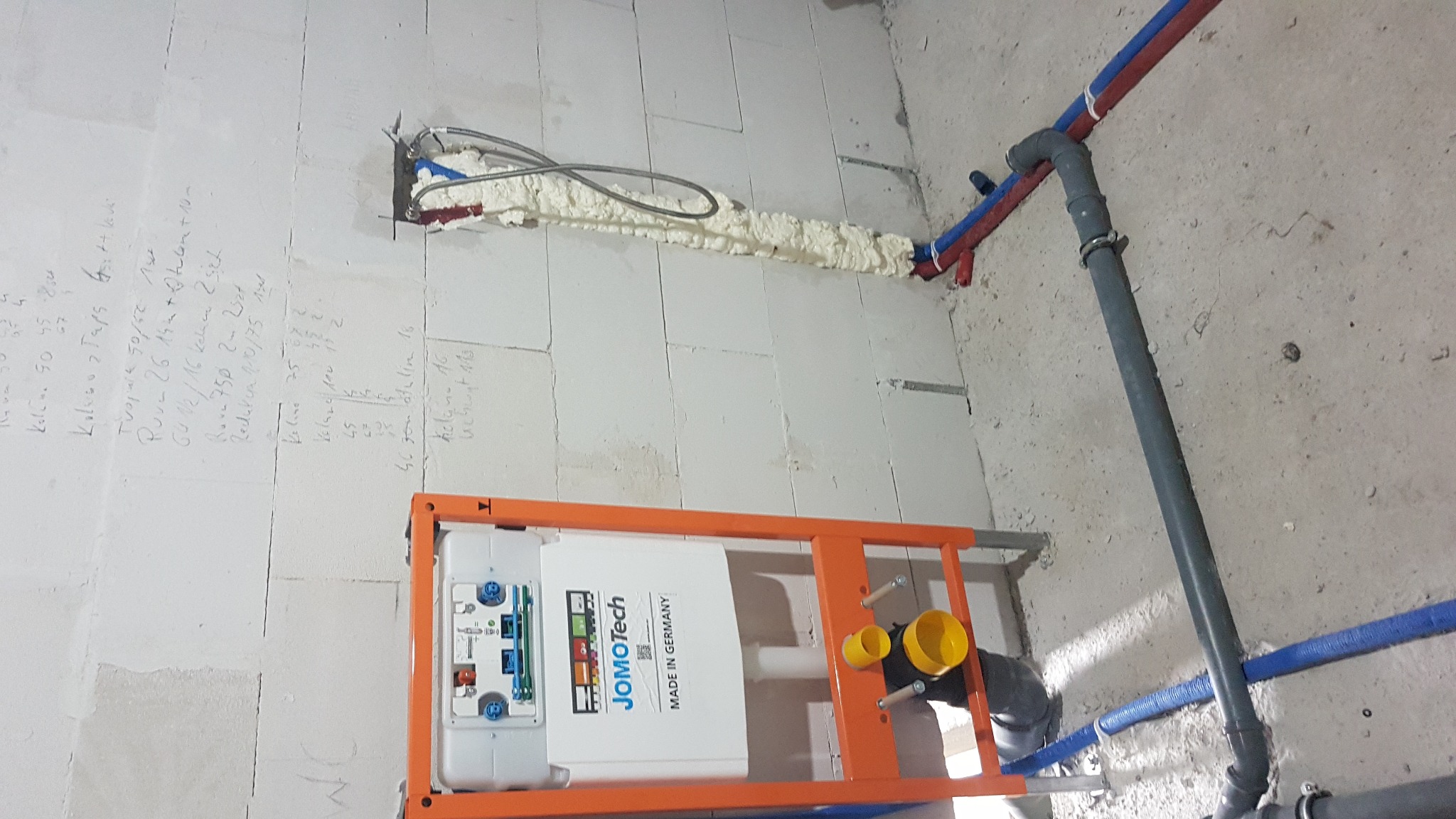 Stelaż podtynkowy WC marki JomoTech, zamontowany w surowym wnętrzu łazienki, widoczne rury instalacji wodnej i kanalizacyjnej, ściana z bloczków z naniesionymi odręcznymi notatkami technicznymi.