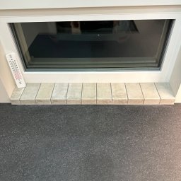 Dawid Filip Filip construction - Wykończony parapet wewnętrzny z jasnych, kwadratowych płytek pod oknem PCV. Obok termometr pokojowy. Szara, matowa podłoga. Ujęcie z dołu, bliski kadr.