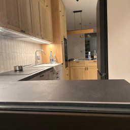 Dawid Filip Filip construction - Jasna, drewniana zabudowa kuchenna z czarnym blatem i płytkami na ścianie, widziana z perspektywy wnętrza. Nowoczesne oświetlenie sufitowe.