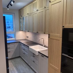 Dawid Filip Filip construction - Jasna kuchnia z drewnianymi szafkami i szarą zabudową, oświetlona reflektorami. Uwagę zwraca ceramiczny zlew farmerski i minimalistyczna armatura.