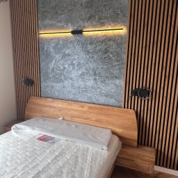 Dawid Filip Filip construction - Nowoczesna sypialnia: drewniane lamele na ścianie, betonowa faktura, podłużna lampa LED nad łóżkiem z drewnianym wezgłowiem i materacem w folii. Minimalistyczny design.