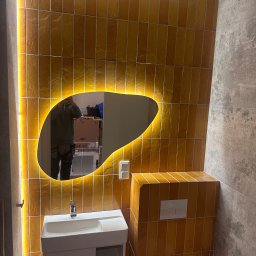 Dawid Filip Filip construction - Żółte, wąskie płytki na ścianie łazienki, podświetlane lustro o nieregularnym kształcie, biała umywalka z szafką i fragment toalety. Nowoczesny design.