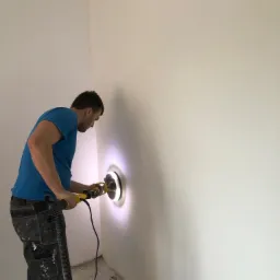 Mężczyzna w niebieskiej koszulce szlifuje ścianę szlifierką oscylacyjną z odkurzaczem, widoczne ślady po szlifowaniu i pył na ścianie.