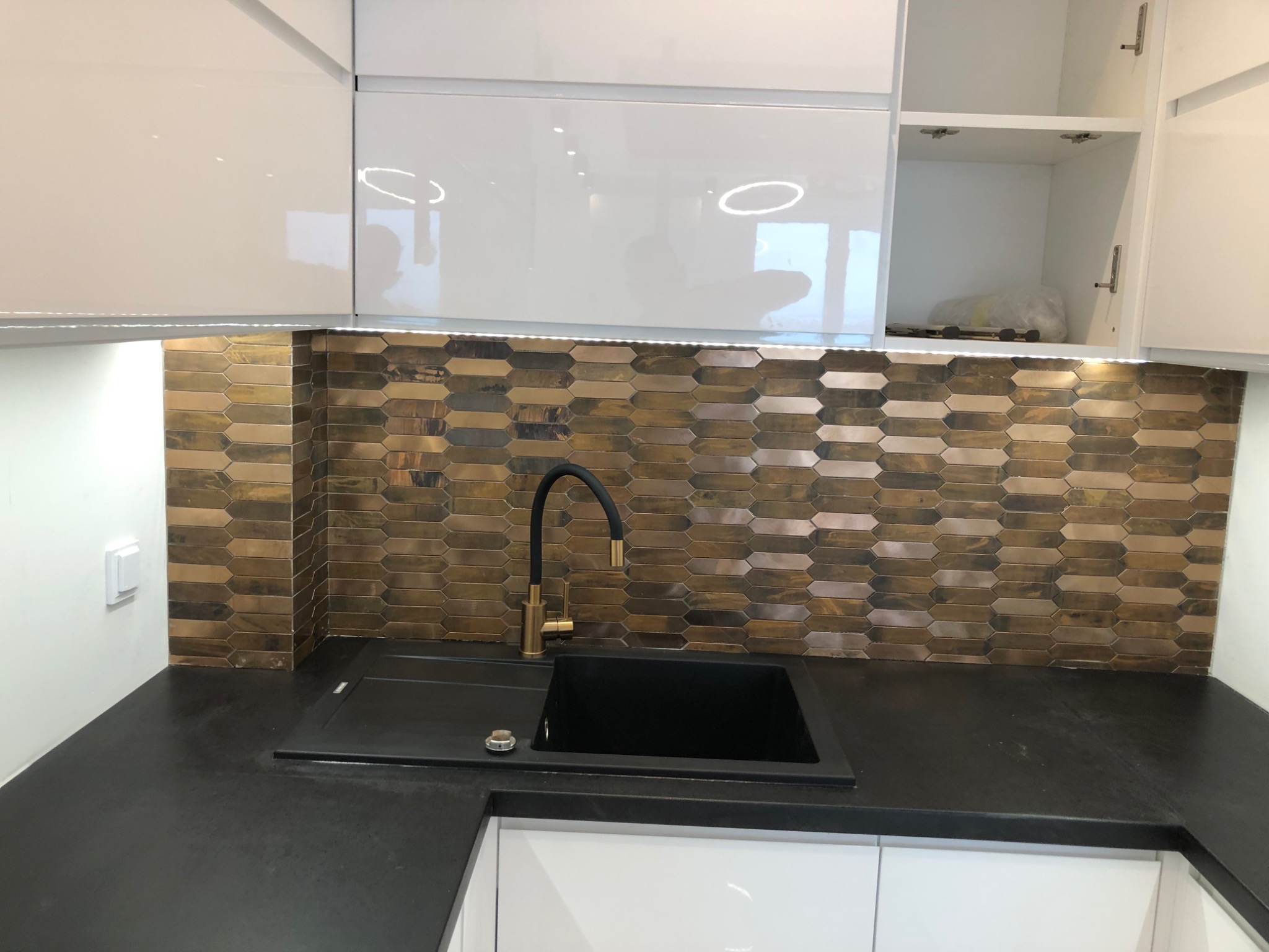 Nowoczesna kuchnia z białymi szafkami, czarnym blatem i zlewozmywakiem oraz mozaiką w odcieniach brązu i złota na ścianie, uzupełniona złotą baterią.