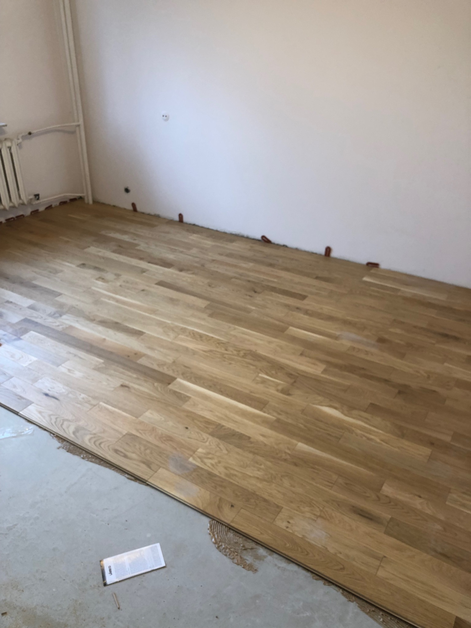 Fragment pokoju z ułożoną częścią jasnej, drewnianej podłogi, widoczne brązowe kliny dystansowe przy ścianie i paczka paneli na podłodze. Podłoże pod panele częściowo przygotowane, widoczny klej.