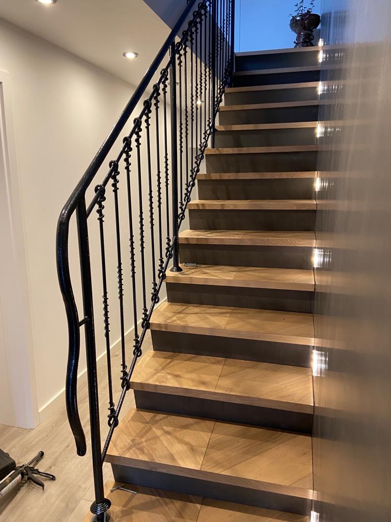Schody z drewnopodobnych płytek ułożonych we wzór jodełki, z czarną, kutą balustradą i oświetleniem LED w stopniach.