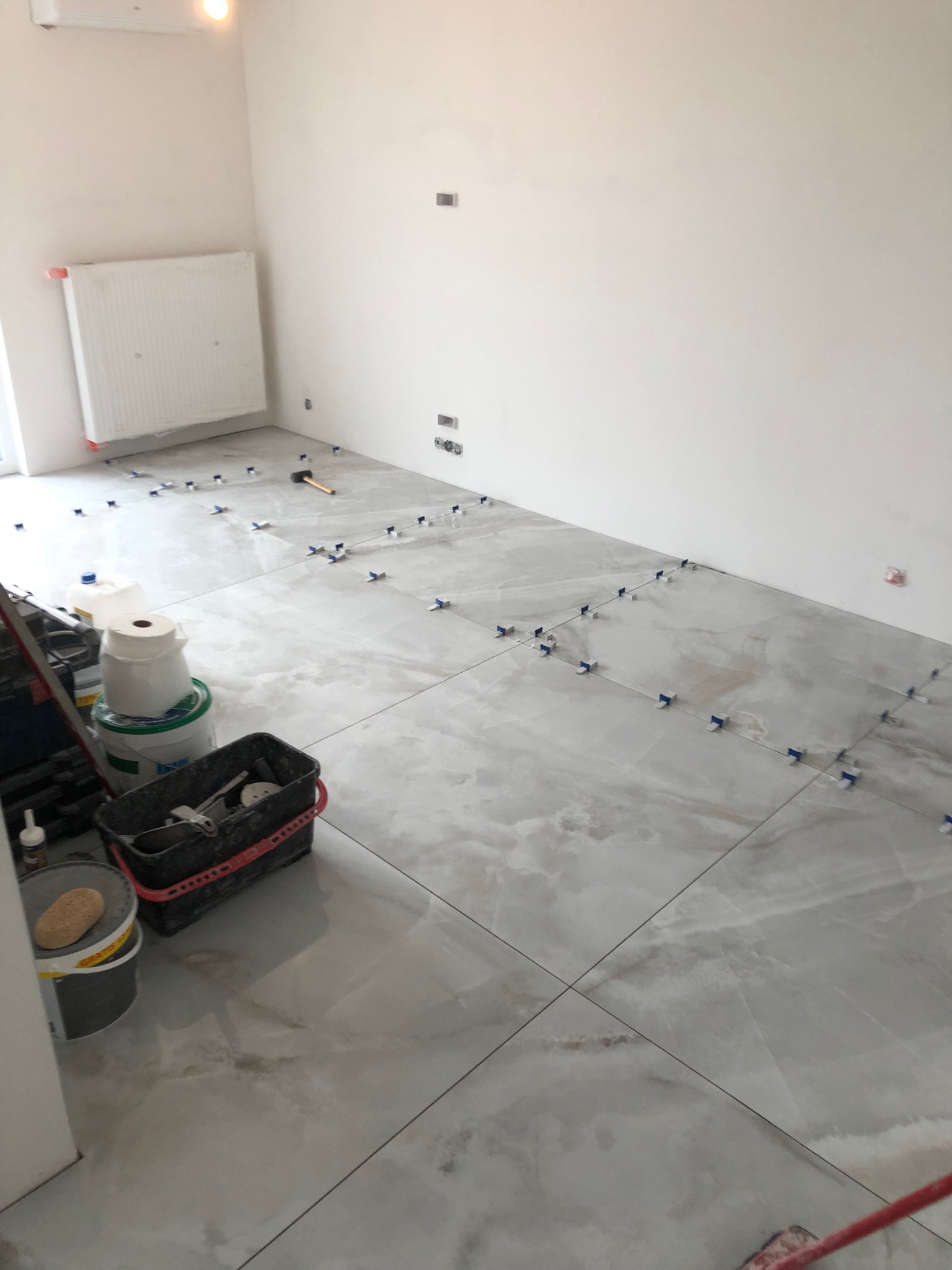 Układanie płytek podłogowych imitujących marmur w jasnym pomieszczeniu; widoczne krzyżyki dystansowe, gumowy młotek i wiadro z narzędziami glazurniczymi.