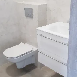 Nowoczesna łazienka z podwieszaną toaletą, białą szafką z umywalką i szarą płytką imitującą kamień na ścianach i podłodze.