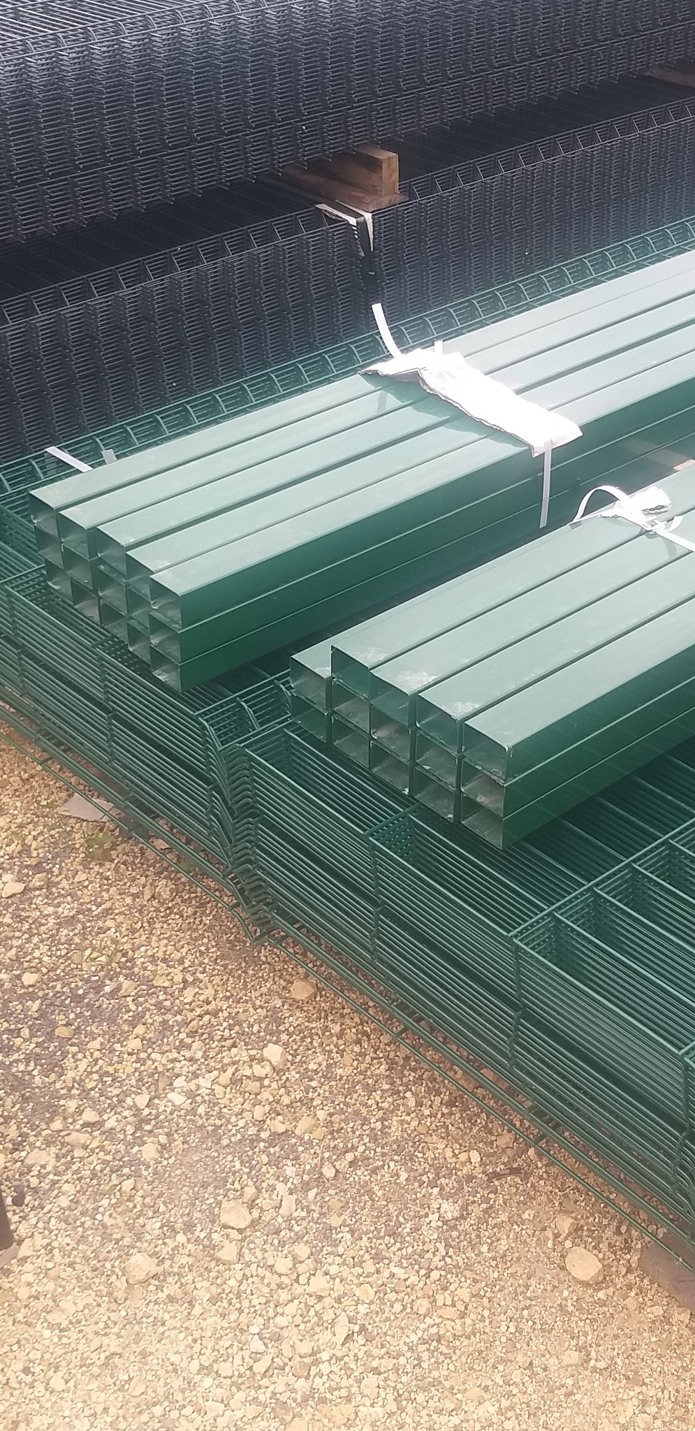 Stos zielonych, kwadratowych słupków ogrodzeniowych i paneli zgrzewanych oczekujących na montaż, widoczne wiązania transportowe.