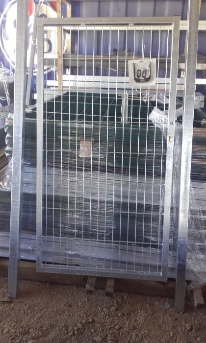 Srebrna, metalowa furtka panelowa z kratką w tle stos paneli ogrodzeniowych zabezpieczonych folią.