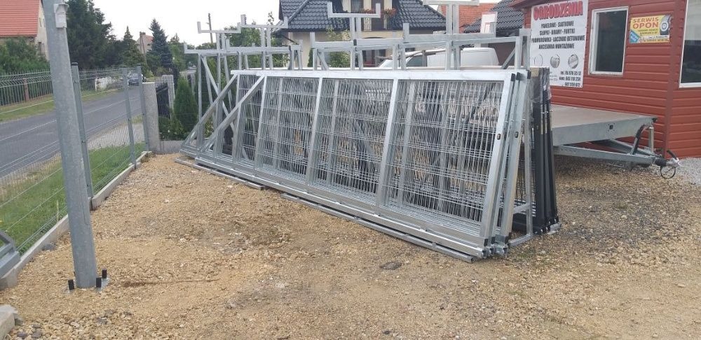 Stos paneli ogrodzeniowych z metalowej siatki, przygotowanych do montażu, na żwirowym podłożu, widoczny fragment zamontowanego ogrodzenia w tle.