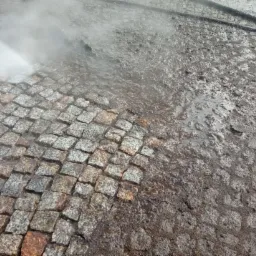 Strumień wody pod ciśnieniem czyści zabrudzoną kostkę brukową, odsłaniając jej naturalny, szaro-pomarańczowy kolor.