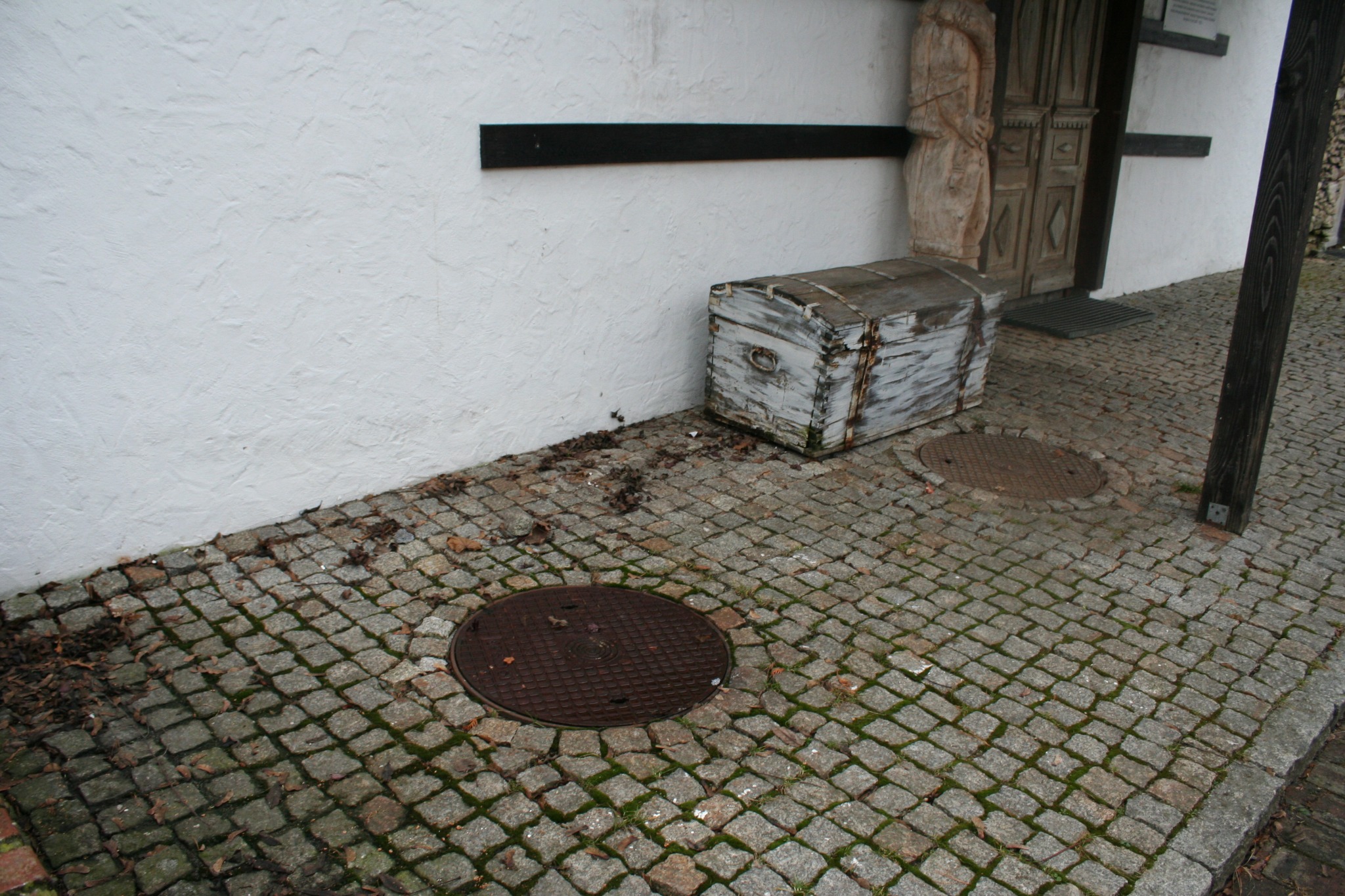 Powierzchnia z kostki brukowej, częściowo pokryta liśćmi, z widocznymi studzienkami kanalizacyjnymi oraz skrzynią w tle, przylegająca do białej, chropowatej ściany budynku z drewnianymi elementami.