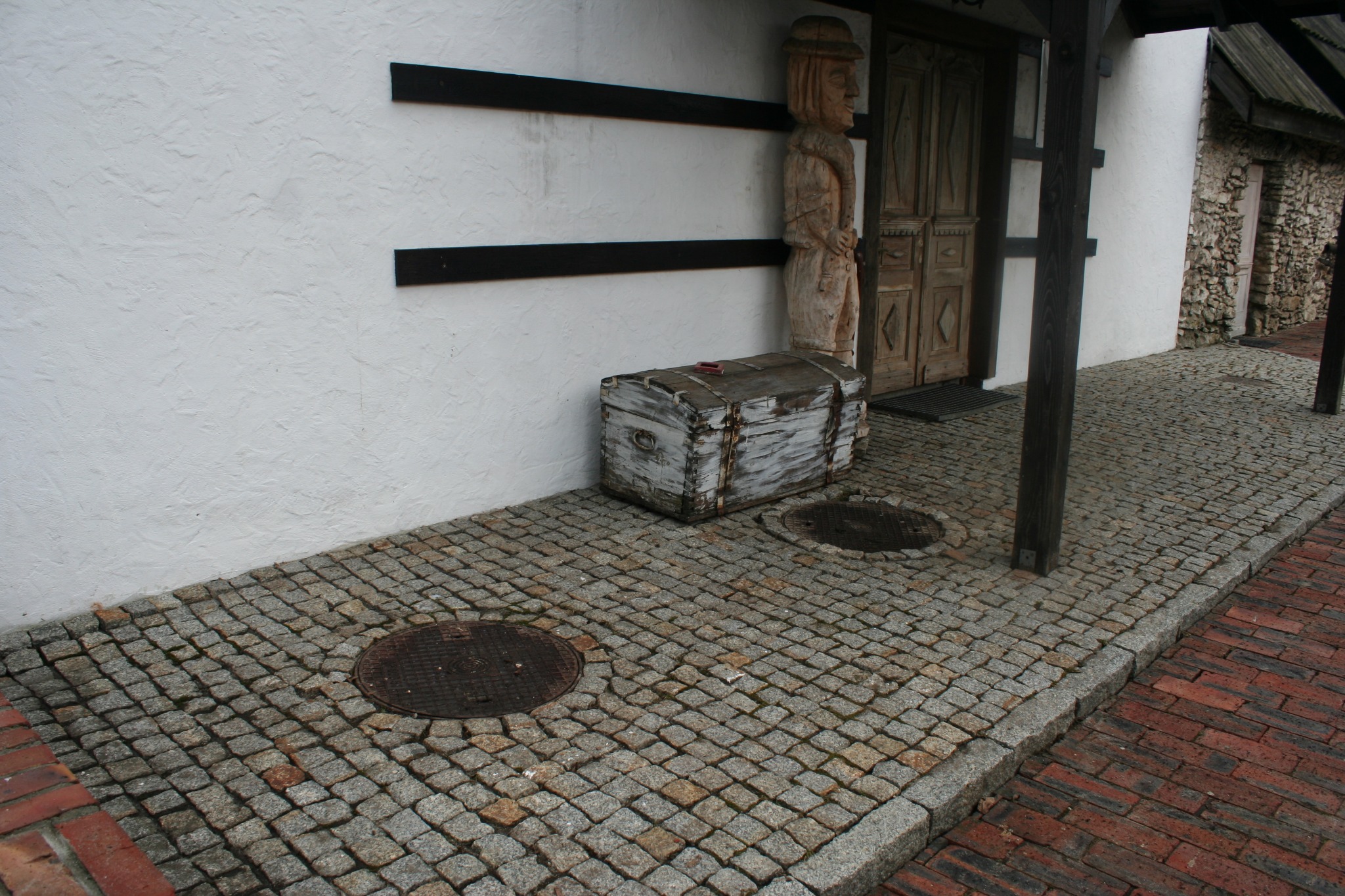 Ułożona kostka brukowa przed wejściem do budynku z białą elewacją, drewnianą rzeźbą i skrzynią, widoczne dwie studzienki kanalizacyjne.