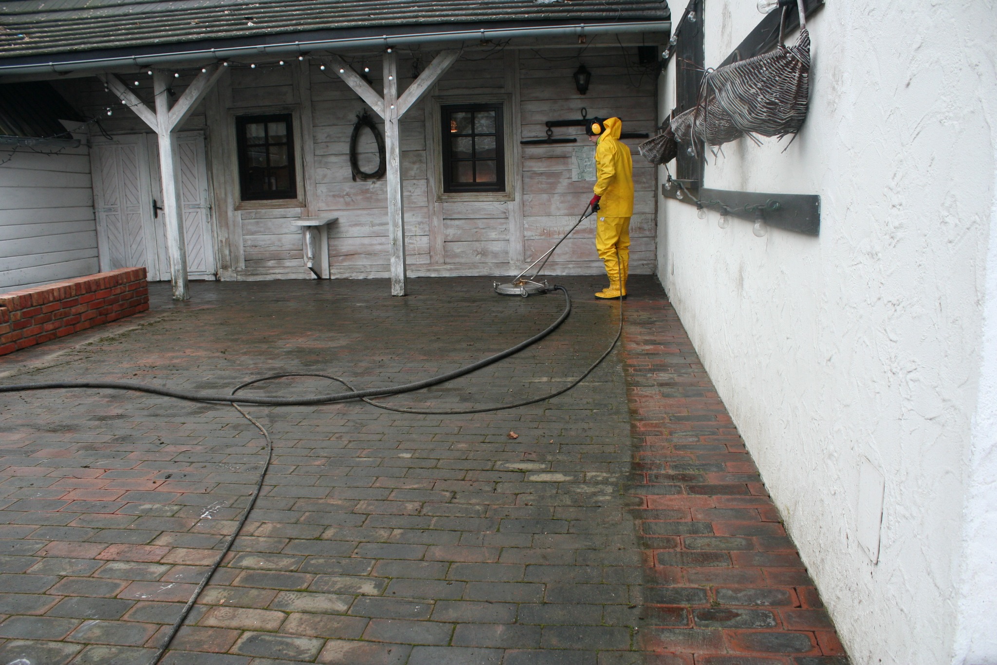 Pracownik w żółtym kombinezonie ochronnym czyści kostkę brukową za pomocą myjki ciśnieniowej z okrągłą dyszą; widoczna różnica w czystości kostki przed i po czyszczeniu, na tle białej ściany...