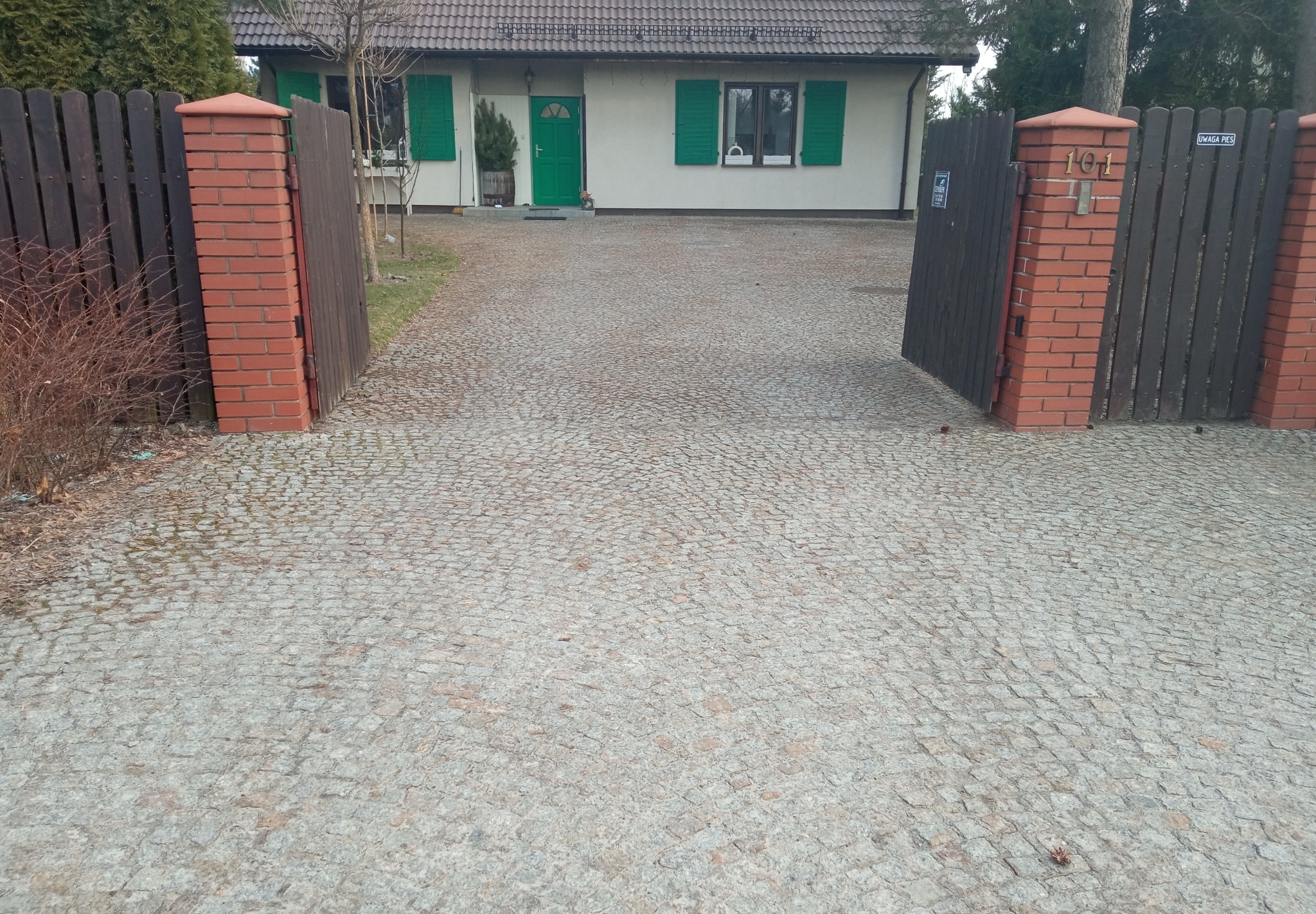 Podjazd do domu wyłożony kostką brukową, brama z ceglanymi słupkami, dom z zielonymi okiennicami w tle.