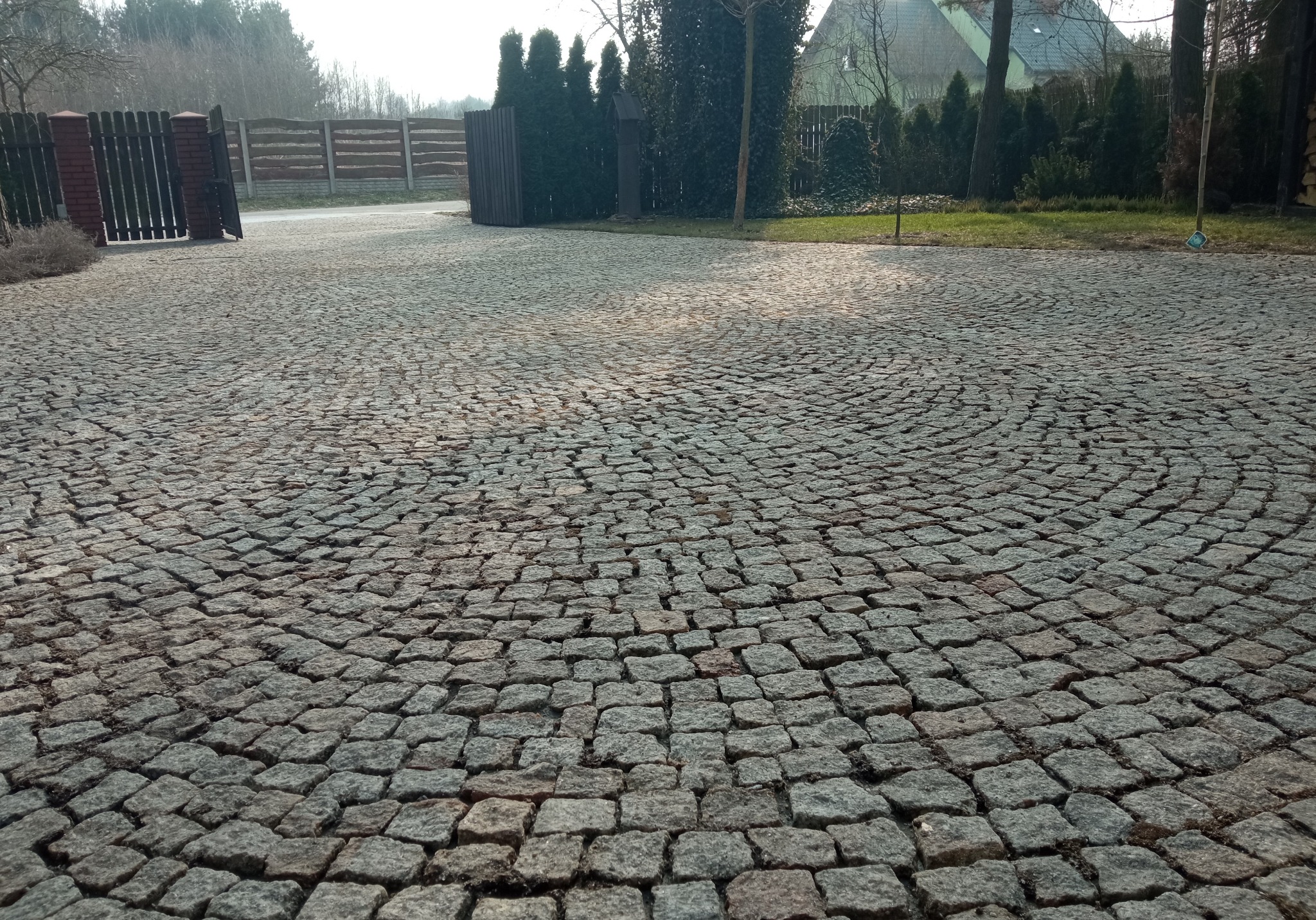 Ułożona w okrąg szara kostka brukowa na podjeździe, widoczne brama wjazdowa i zieleń w tle.