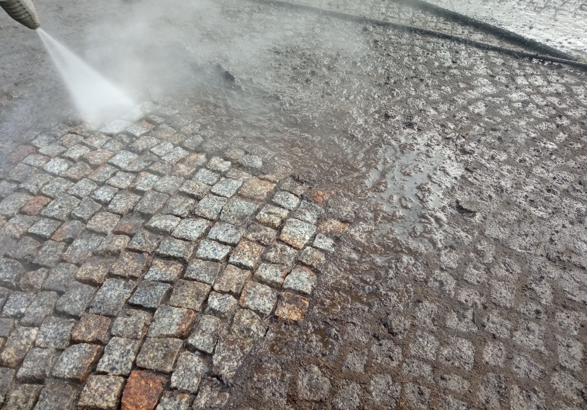 Strumień wody pod ciśnieniem czyści zabrudzoną kostkę brukową, odsłaniając jej naturalny, szaro-pomarańczowy kolor.