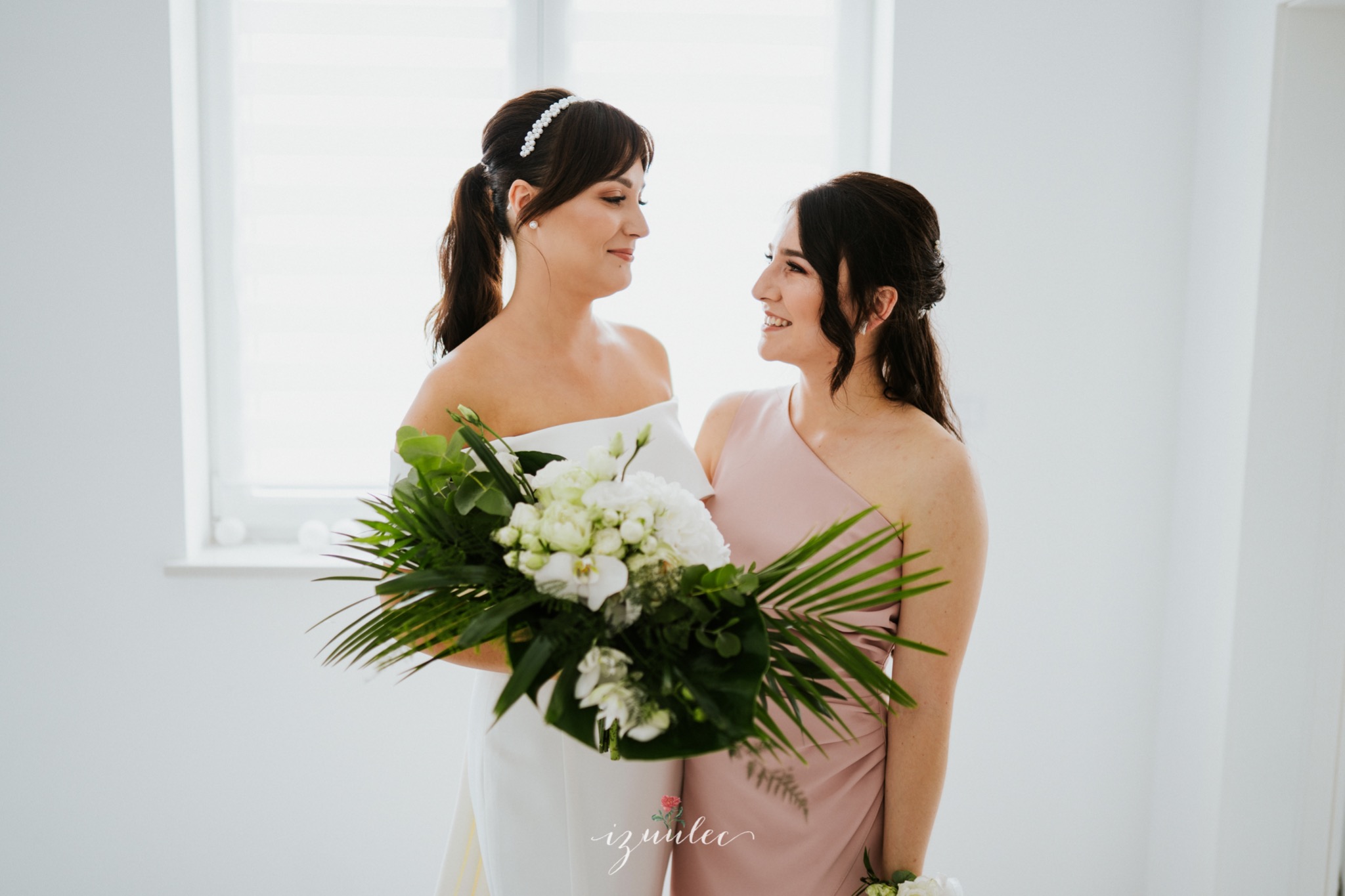 Panna młoda w białej sukni i druhna w różowej sukni patrzą na siebie z uśmiechem, panna młoda trzyma bukiet białych kwiatów i zielonych liści na jasnym tle.