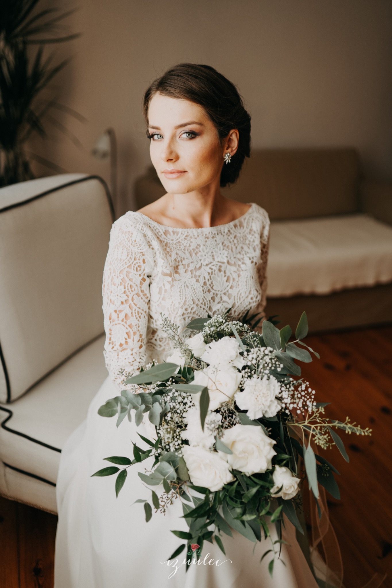 Panna młoda w koronkowej sukni z bukietem białych róż i eukaliptusa, siedząca na krześle w jasnym wnętrzu.