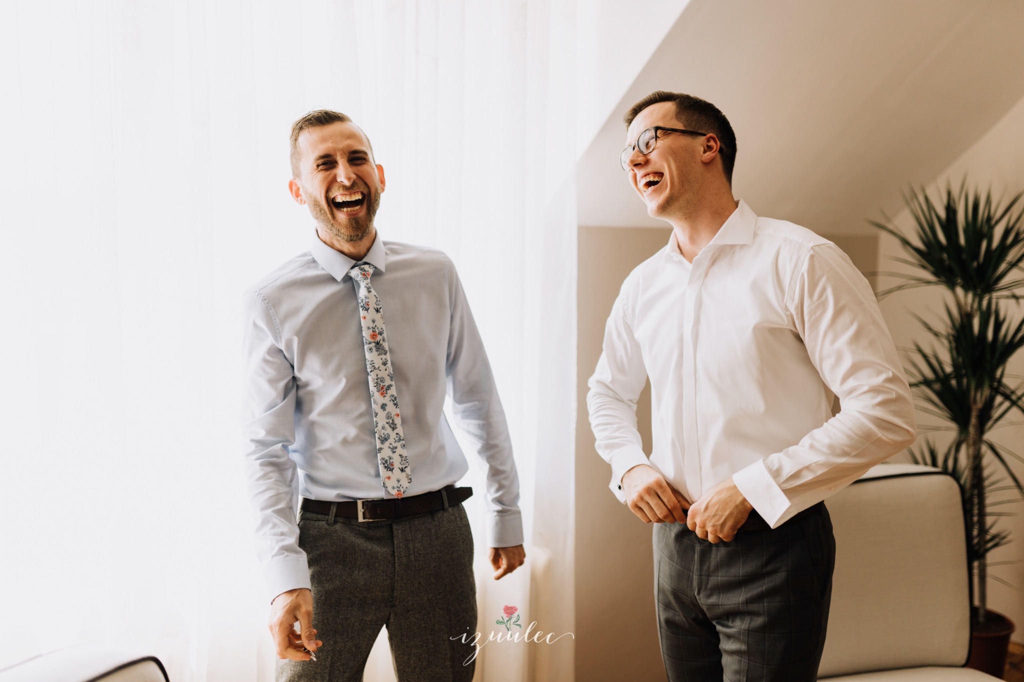 Dwóch roześmianych mężczyzn w koszulach i spodniach, jeden z krawatem w kwiaty, przygotowujących się do uroczystości, sfotografowanych w jasnym wnętrzu.