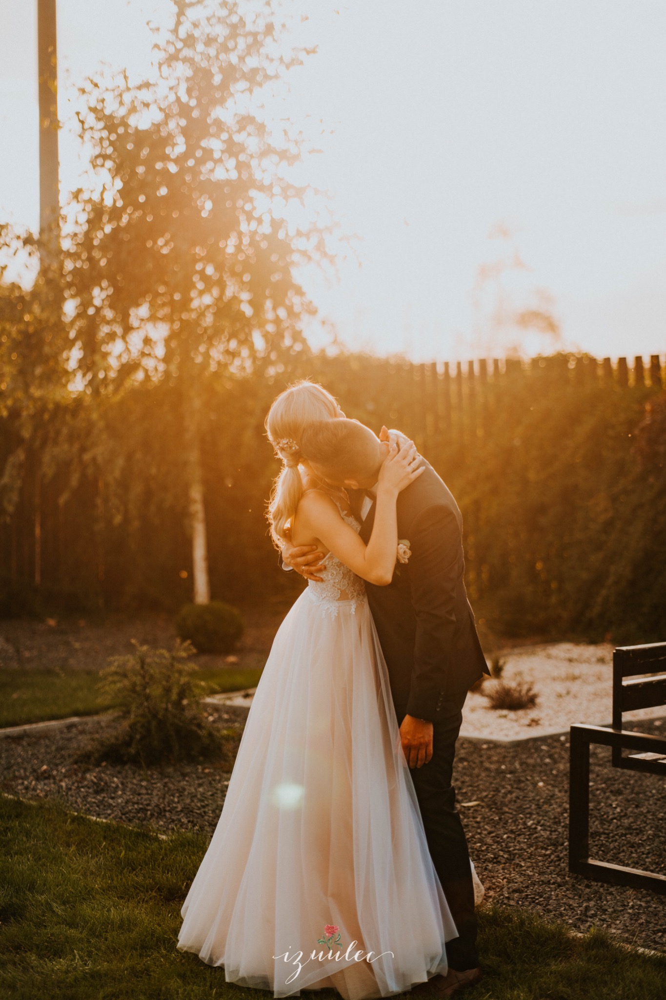 Para młoda w objęciach na tle zachodzącego słońca, panna młoda w zwiewnej sukni z koronkową górą, pan młody w ciemnym garniturze, w tle drzewa i ławka.