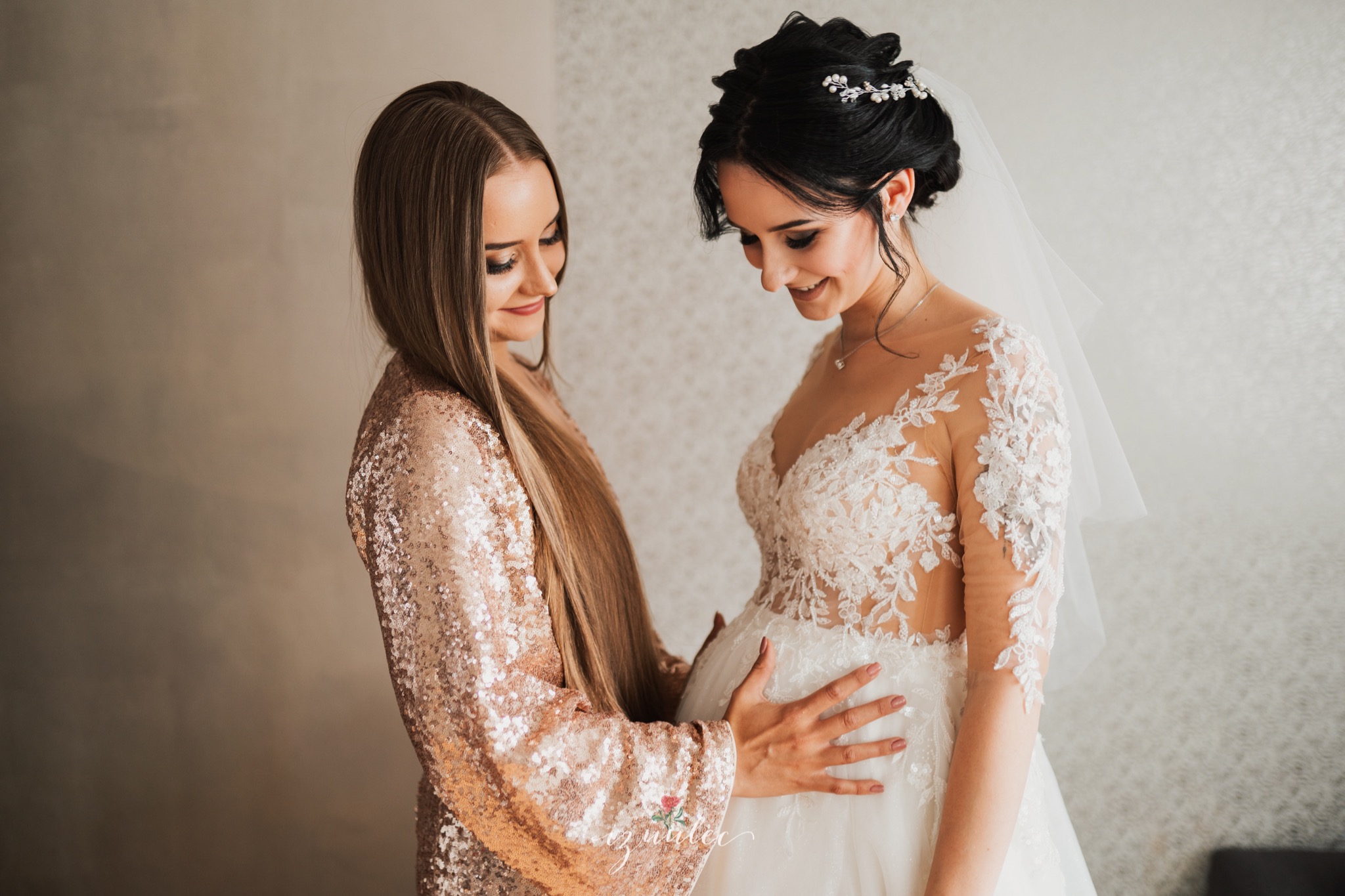 Panna młoda w koronkowej sukni ślubnej z welonem, obejmowana przez druhnę w cekinowej sukience, obie z uśmiechem patrzą w dół.