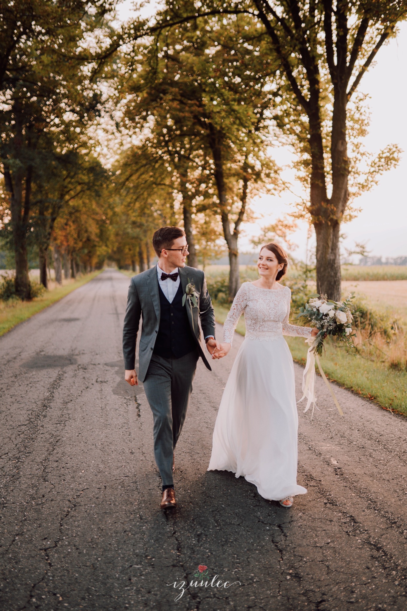 Para młoda spaceruje po wiejskiej drodze w promieniach zachodzącego słońca; panna młoda w koronkowej sukni z bukietem, pan młody w szarym garniturze i muszce.