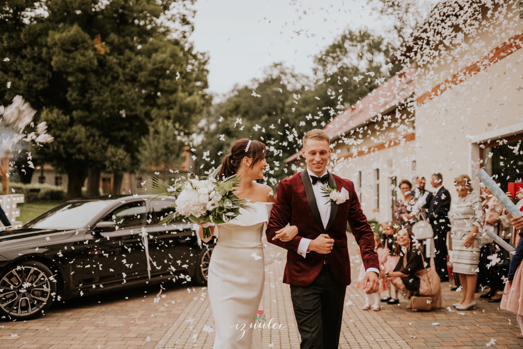 Uśmiechnięta para młoda, otoczona konfetti, idzie po brukowanej ścieżce. Panna młoda w białej sukni z bukietem, pan młody w bordowym smokingu. W tle czarny samochód i goście.