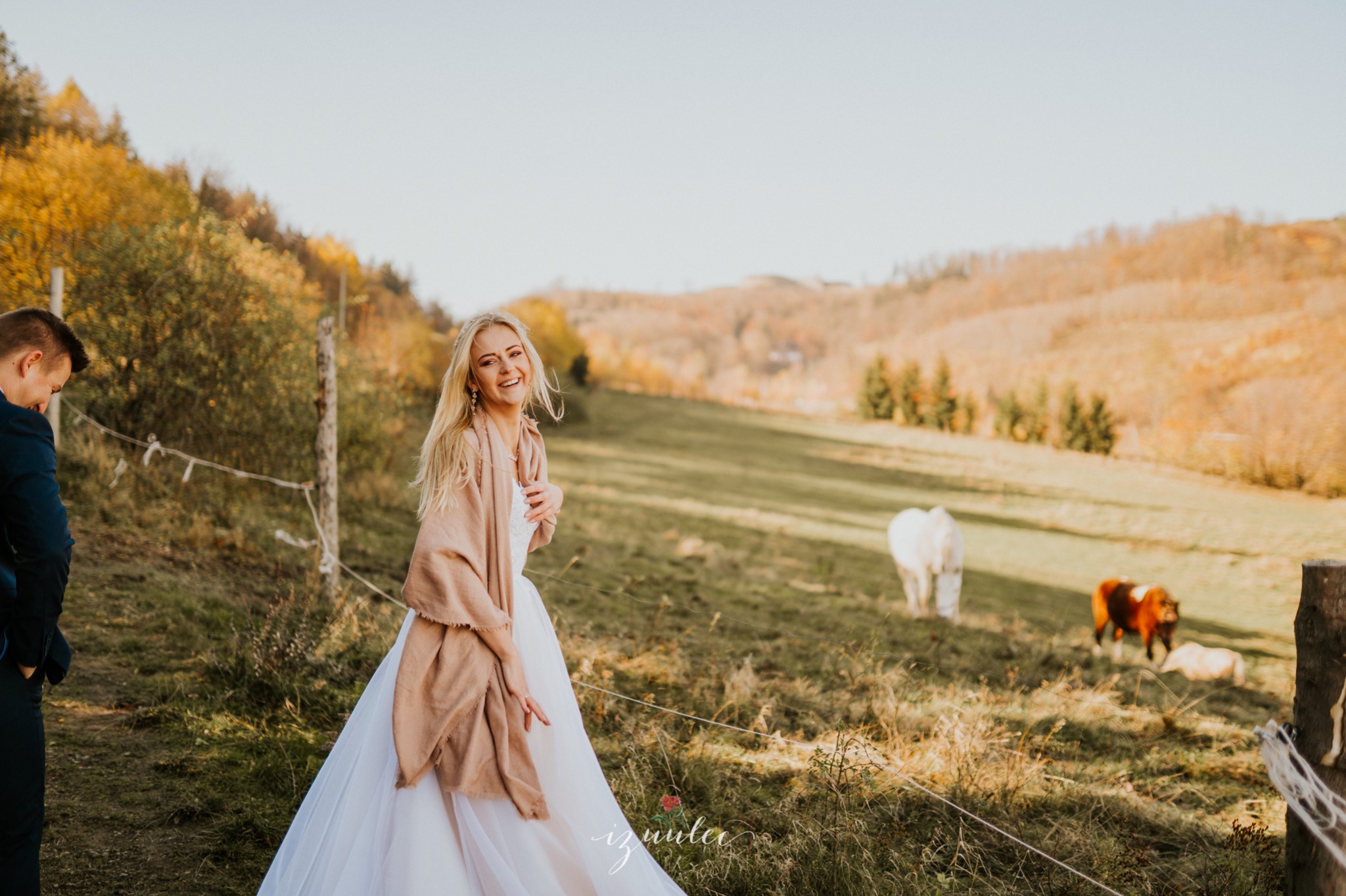 Uśmiechnięta panna młoda w białej sukni i beżowym szalu pozuje na tle jesiennego krajobrazu z końmi na pastwisku, obok stoi pan młody w granatowym garniturze.