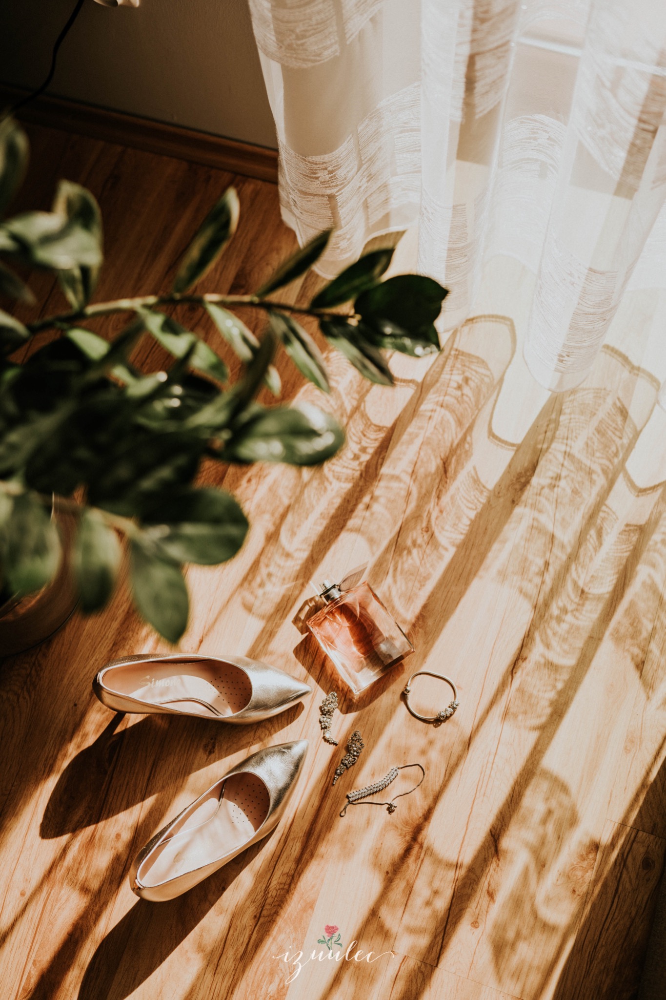 Złote baleriny, flakon perfum, bransoletka i kolczyki leżące na drewnianej podłodze w promieniach słońca wpadających przez firankę, obok doniczka z zieloną rośliną.