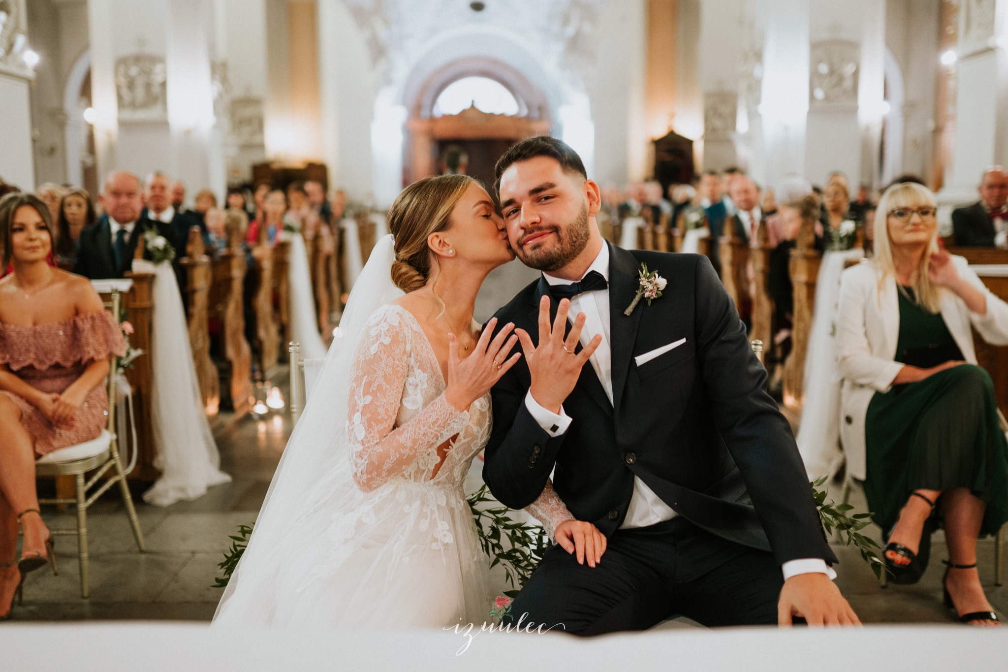 Panna młoda w koronkowej sukni całuje pana młodego w czarnym smokingu, oboje prezentują obrączki na tle kościoła z gośćmi.