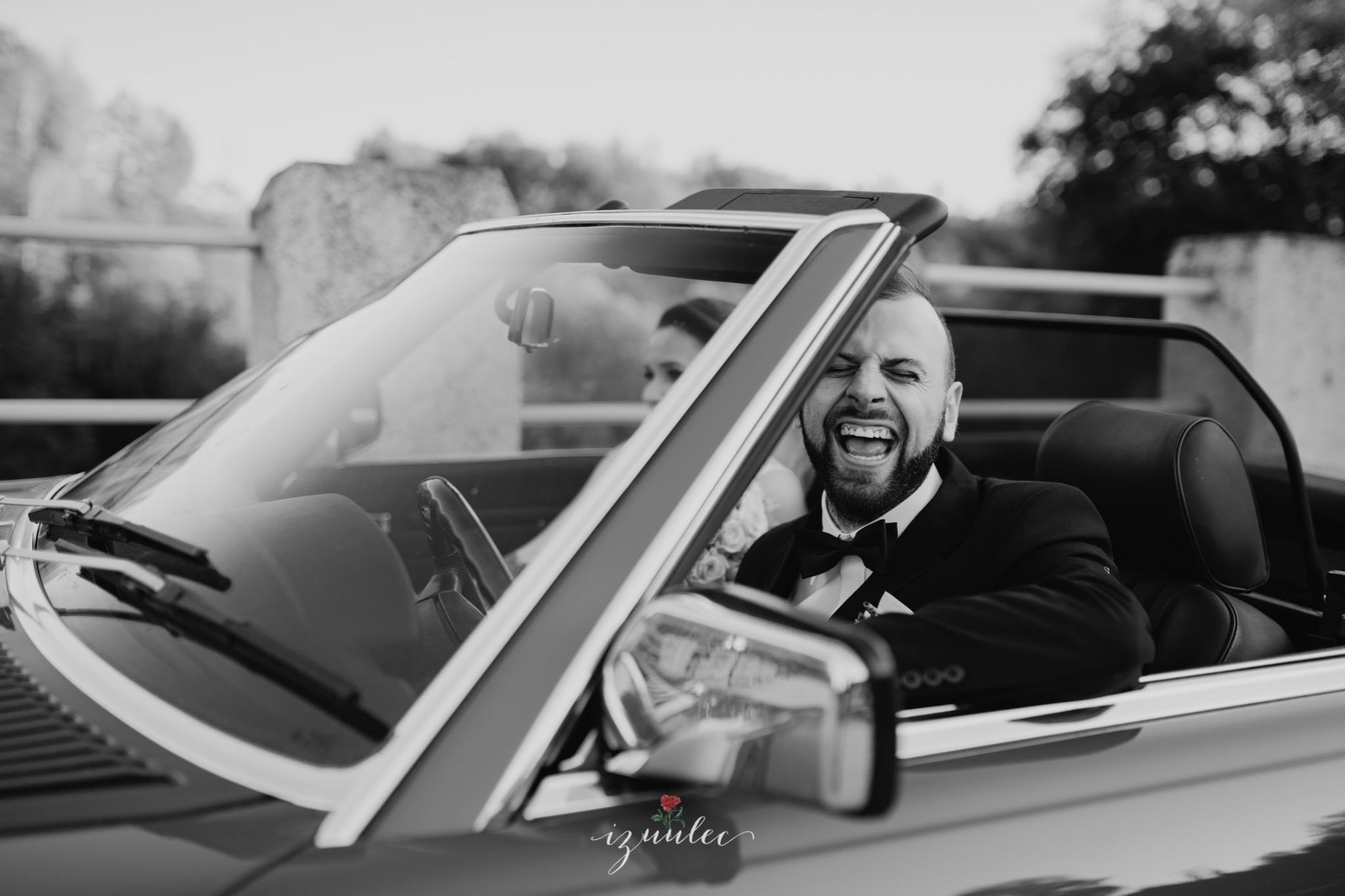 Czarno-białe zdjęcie pana młodego w kabriolecie, szeroki uśmiech, za nim panna młoda, w tle kamienny mur.