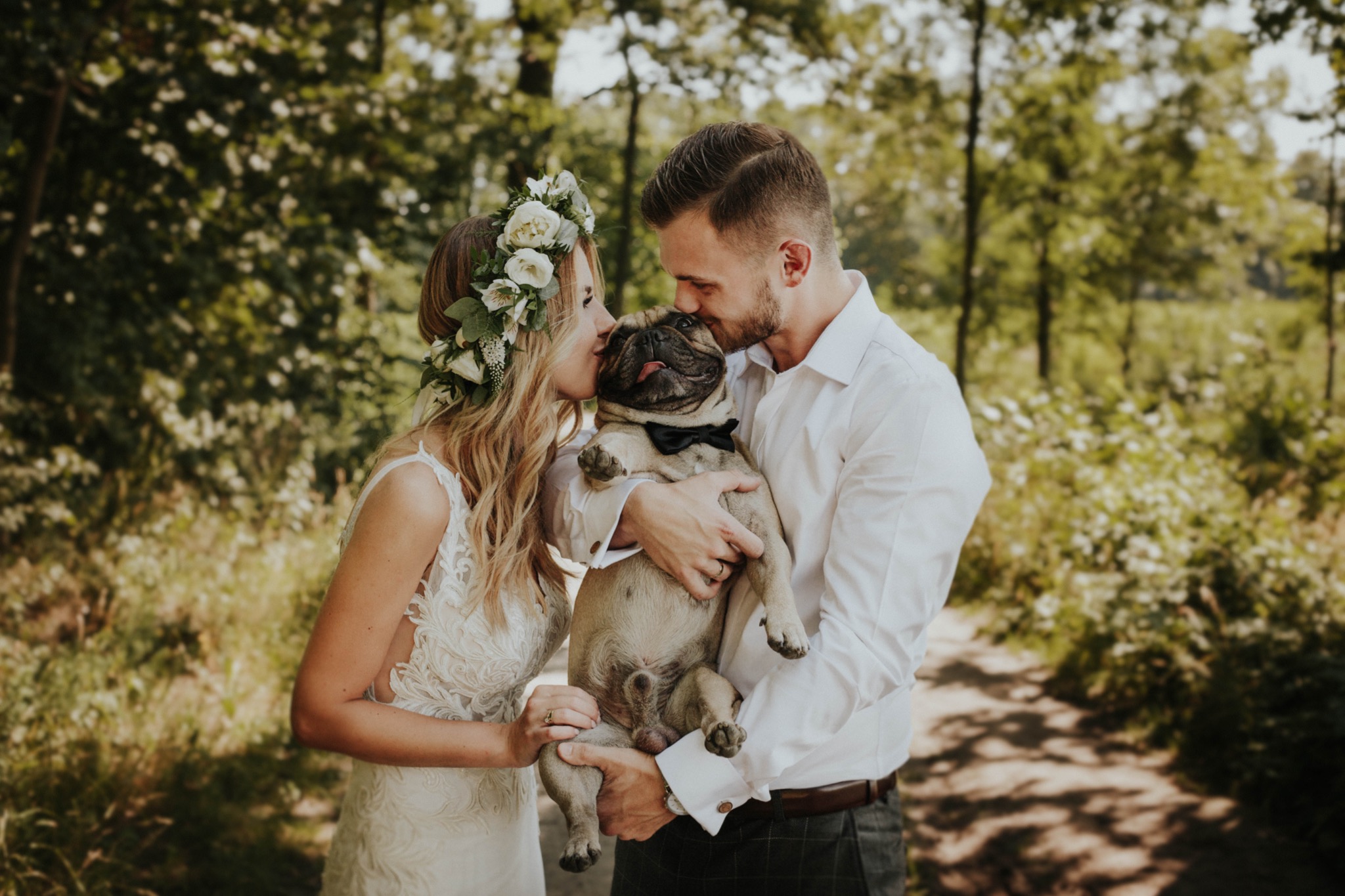 Para młoda w lesie całuje mopsa w czarnej muszce, panna młoda w wianku z białych kwiatów, delikatna suknia ślubna z koronką, pan młody w białej koszuli.