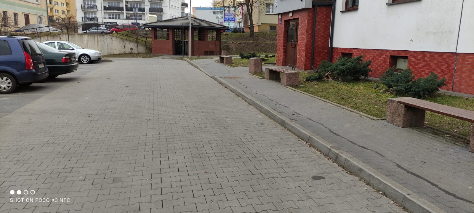 Umyta kostka brukowa na parkingu osiedlowym, widoczna wiata śmietnikowa, zaparkowane samochody i ławki.