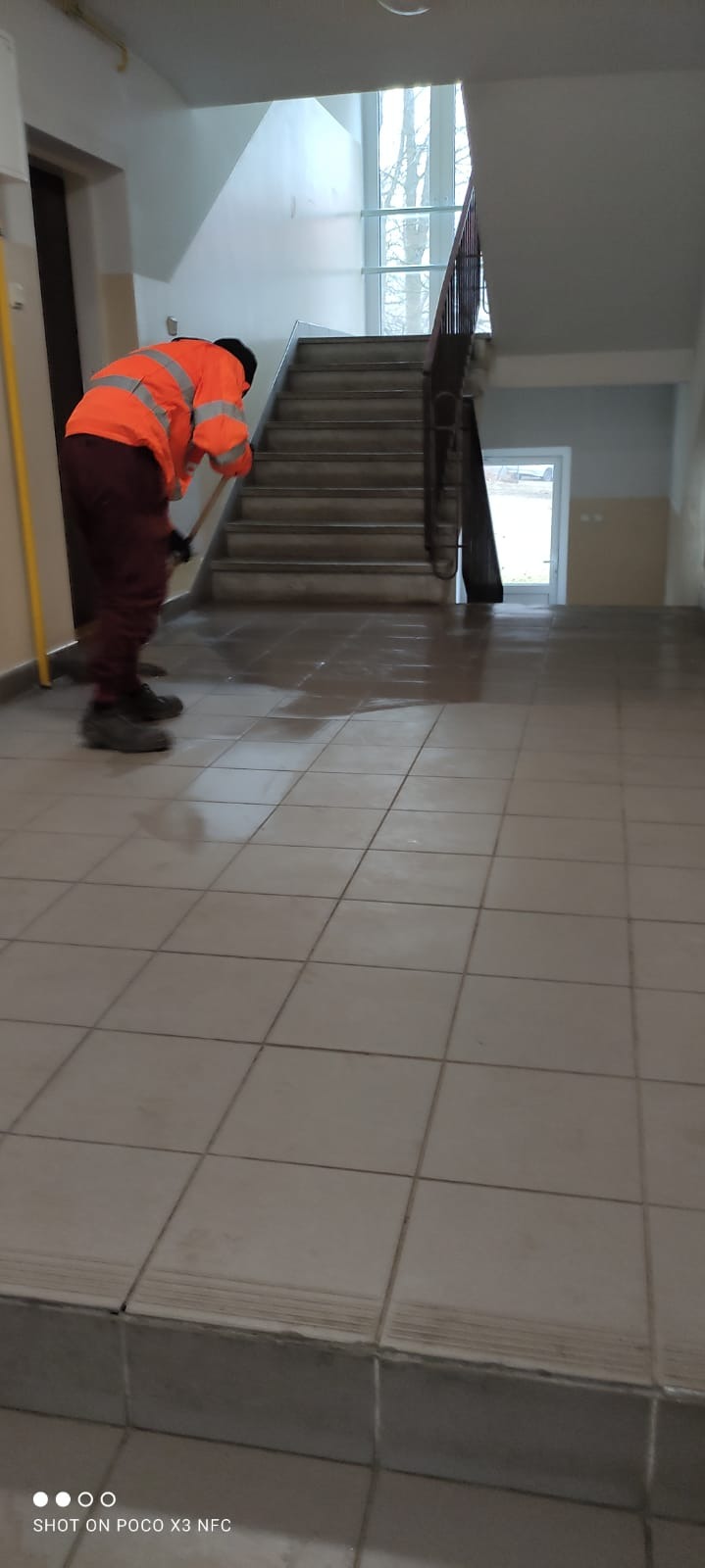 Pracownik w pomarańczowej kamizelce odblaskowej zamiata mokrą podłogę w holu budynku z widocznymi schodami i drzwiami.