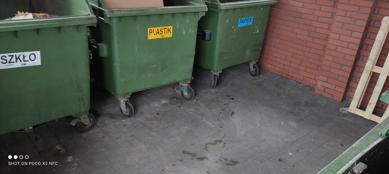 Rząd zielonych kontenerów na odpady: szkło z zawartością, plastik z kartonem i papier, ustawione przy ceglanym murze i palecie.