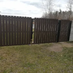 Nowoczesna, brązowa brama panelowa z metalowymi słupkami, częściowo otwarta, widok na zewnątrz posesji, obok betonowe ogrodzenie, trawnik z opadłymi liśćmi, pochmurne niebo.