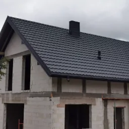 Dom w trakcie budowy z nowym, ciemnoszarym dachem z blachodachówki, widoczny komin, rynna i nieotynkowane ściany z betonu komórkowego.