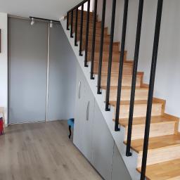 Gregor - Drewniane schody z czarną metalową balustradą i szarą zabudową szafek pod nimi, widoczne przesuwne drzwi szafy wnękowej i panele podłogowe.