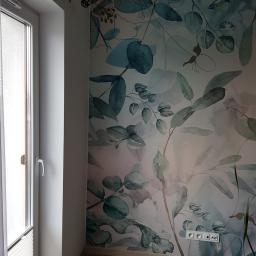 Gregor - Fragment pokoju z tapetą w motywy roślinne, widoczne gniazdka elektryczne, okno z żaluzjami plisowanymi i drewniana podłoga.