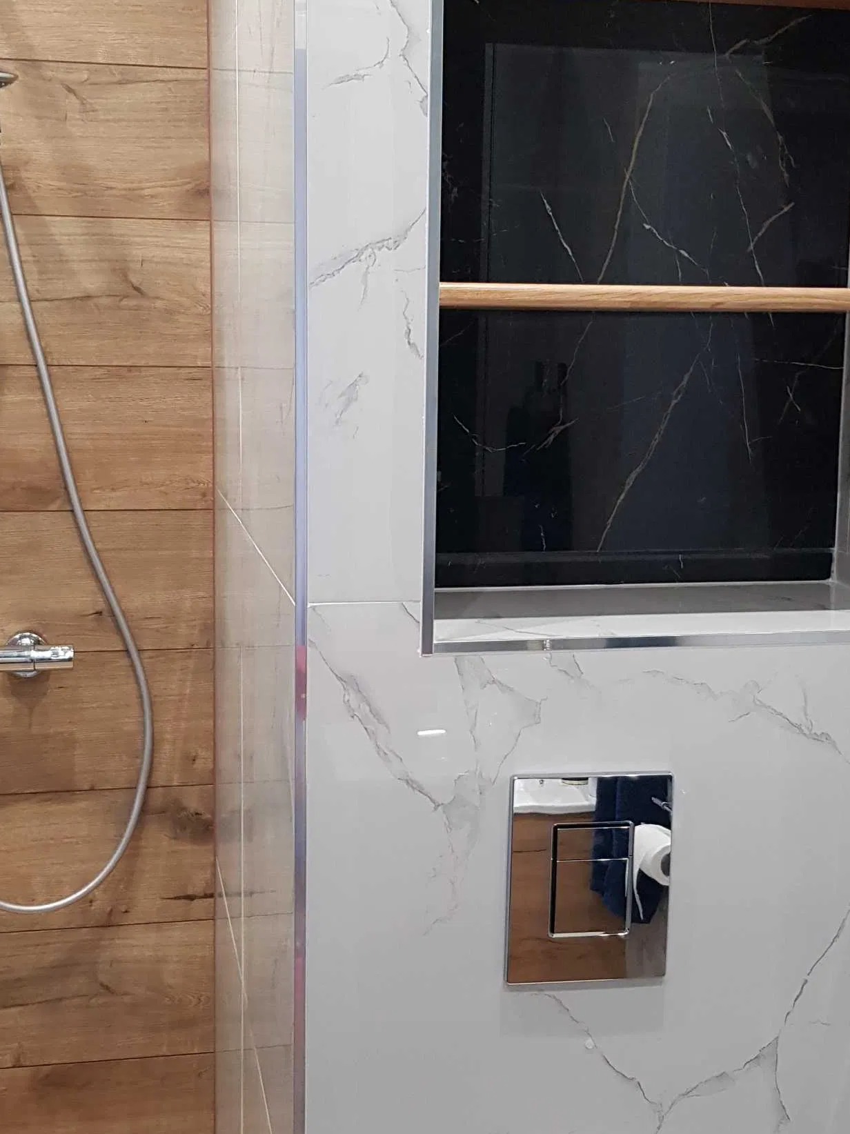 Wykończona łazienka z drewnopodobnymi płytkami, szklaną kabiną prysznicową i zabudowaną półką z czarnym marmurem, widoczny chromowany panel spłukujący.