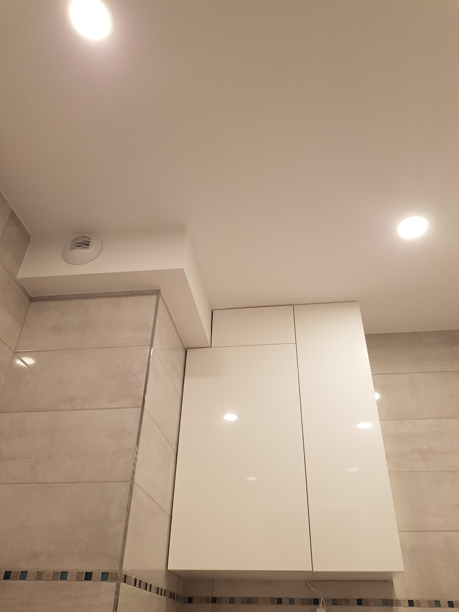 Białe, lakierowane szafki wiszące w łazience, dopasowane do zabudowy sufitowej z kratką wentylacyjną, beżowe płytki na ścianach z dekoracyjnym pasem, oświetlenie punktowe w suficie.