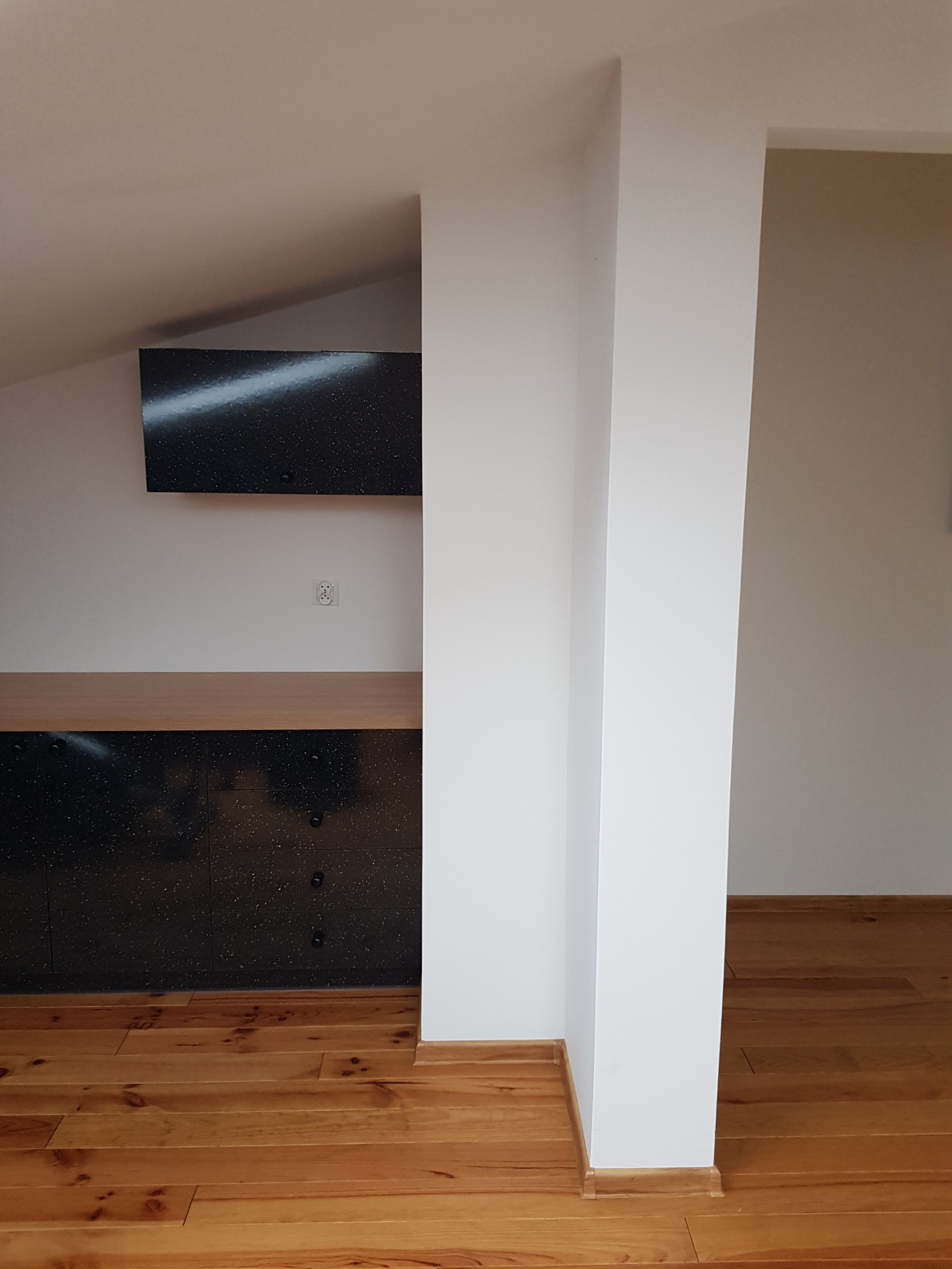 Część pokoju na poddaszu z drewnianą podłogą, zabudową meblową z czarnymi frontami z drobnymi białymi plamkami i drewnianym blatem, biała ściana z gniazdkiem elektrycznym i biały filar