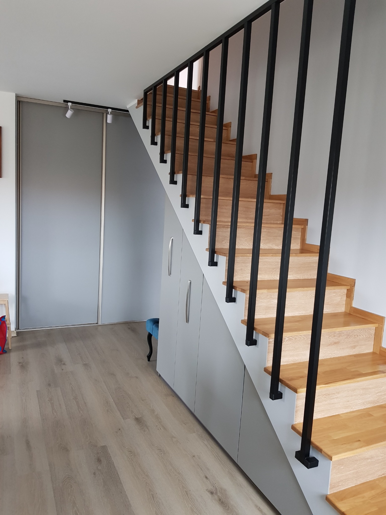 Drewniane schody z czarną metalową balustradą i szarą zabudową szafek pod nimi, widoczne przesuwne drzwi szafy wnękowej i panele podłogowe.