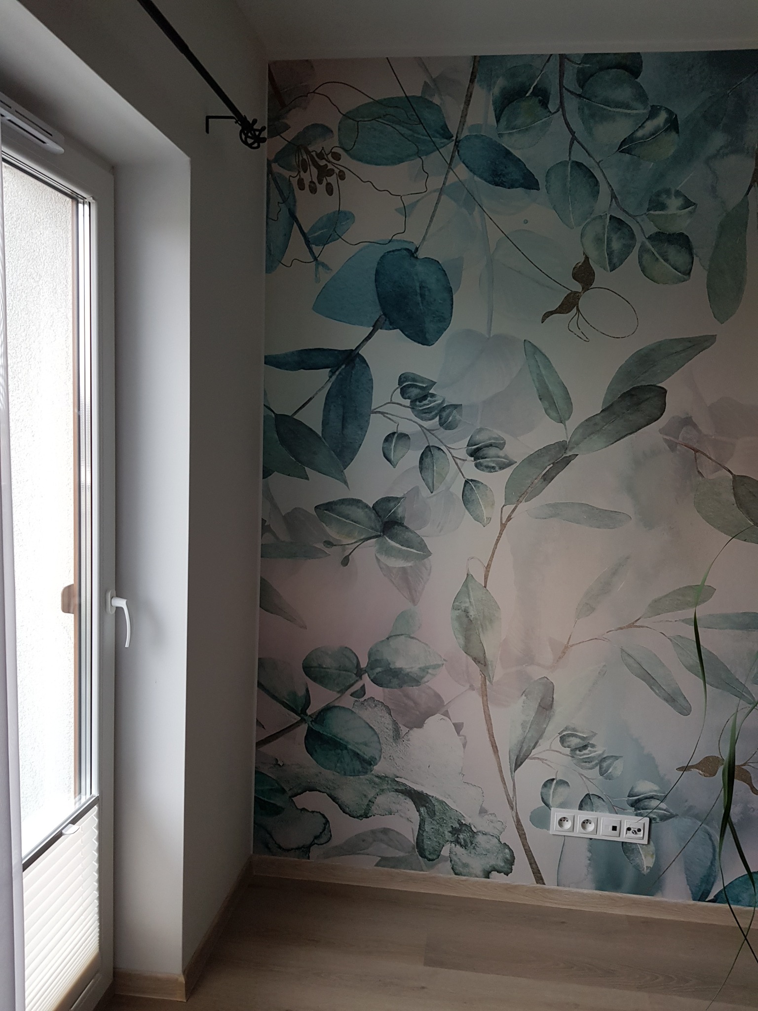 Fragment pokoju z tapetą w motywy roślinne, widoczne gniazdka elektryczne, okno z żaluzjami plisowanymi i drewniana podłoga.