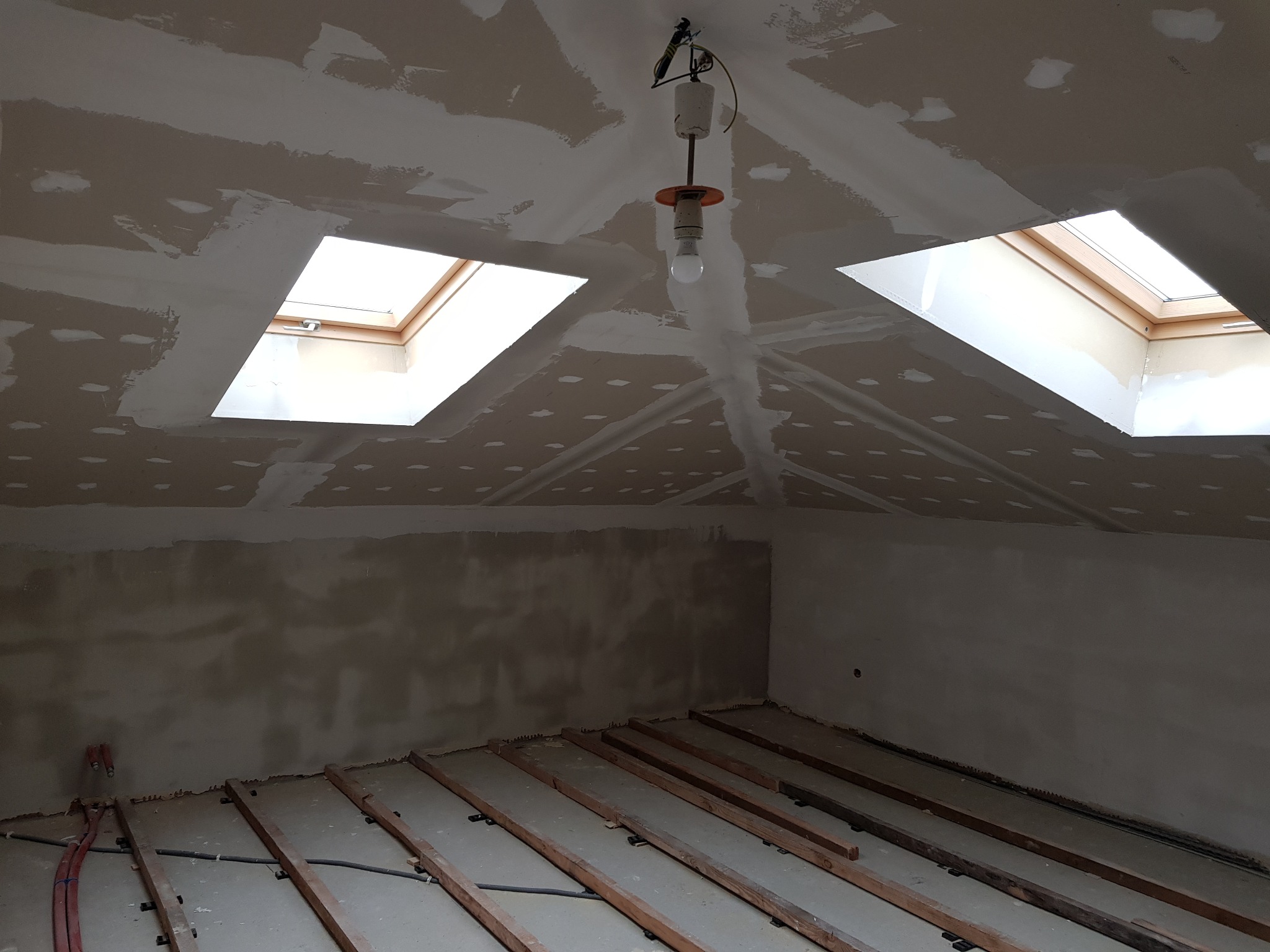 Poddasze w trakcie prac wykończeniowych: surowy sufit z dwoma oknami dachowymi, prowizoryczne oświetlenie, przygotowany podkład pod podłogę z widocznymi elementami instalacji grzewczej.