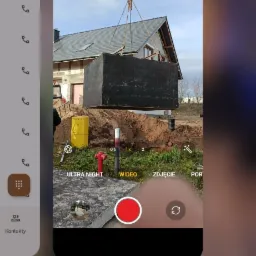Ekran smartfona z otwartą aplikacją aparatu, na której widoczny jest czarny, betonowy zbiornik zawieszony na łańcuchach dźwigu, w tle wykop i budynek w trakcie budowy, widoczny czerwony hydrant