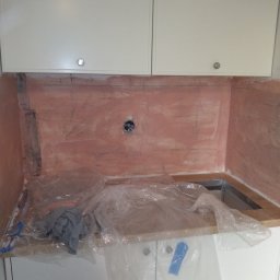 Usługi Remontowo Budowlane - Przygotowanie ściany pod płytki w kuchni: nałożona warstwa gruntująca w kolorze różowym, widoczne ślady po ołówku, zabezpieczone folią meble kuchenne.